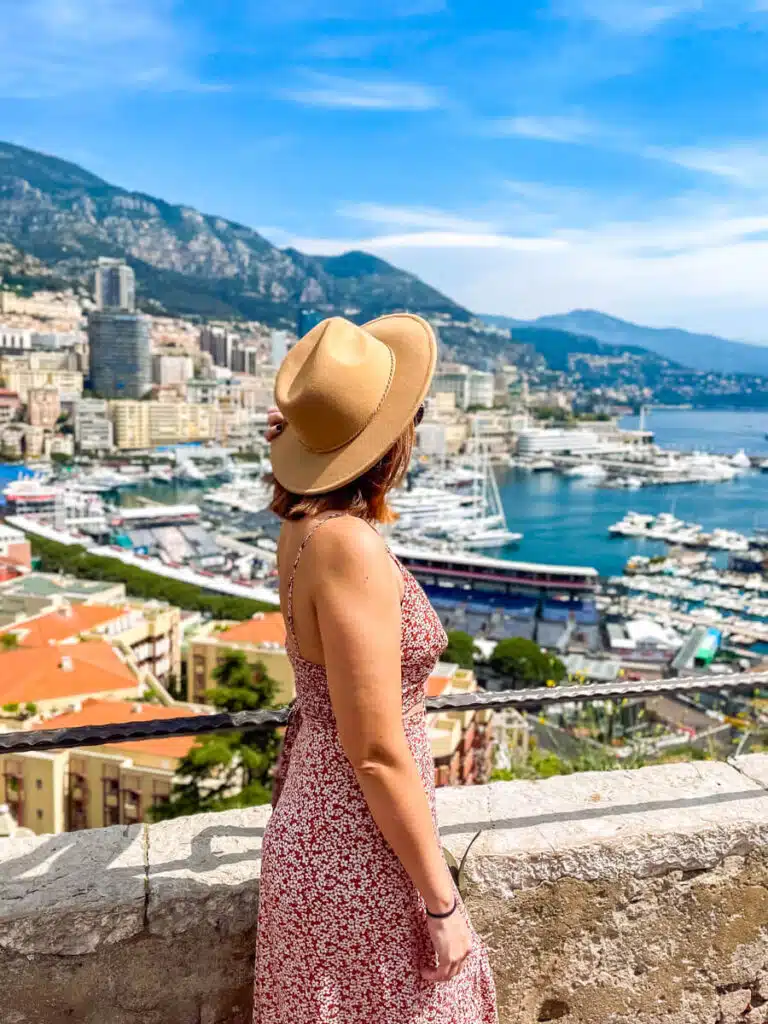 A woman in a sunhat and floral dress gazes over the harbor of Monaco, with luxury yachts and terracotta rooftops below during a day trip to Monaco from Nice.