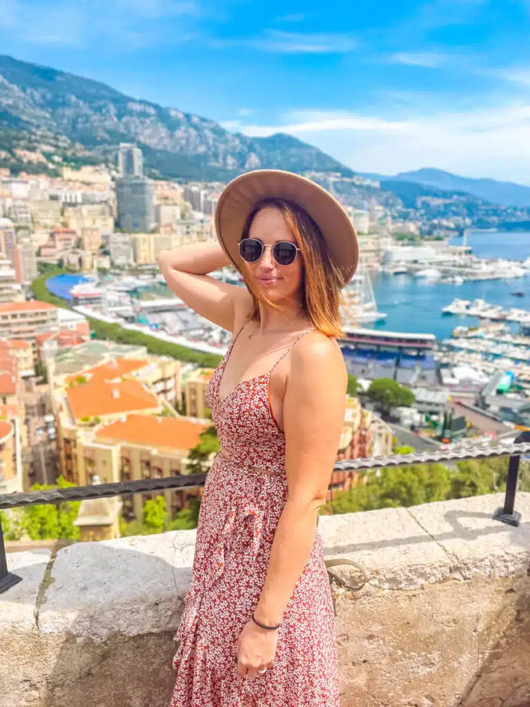 A woman in a hat and sunglasses poses with the scenic port of Monaco behind her, surrounded by mountains and high-rise buildings on a day trip from Nice, France.