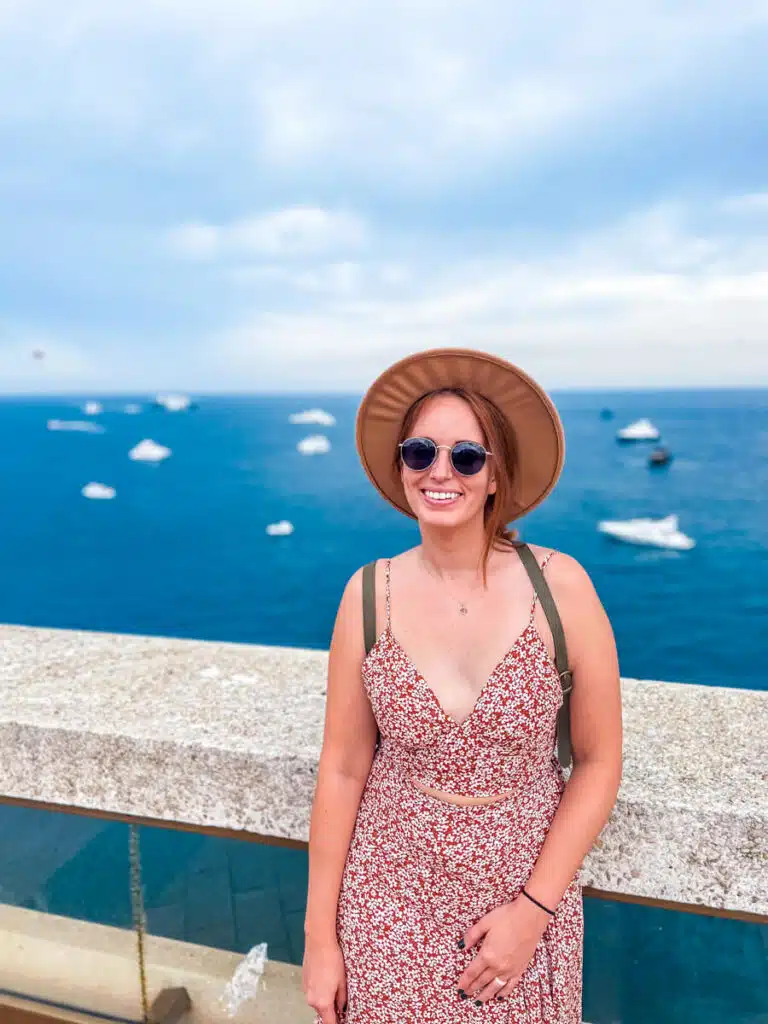 A smiling woman in a red floral dress and hat stands by the sea in Monaco, with dozens of yachts dotting the water behind her during a day trip from Nice.