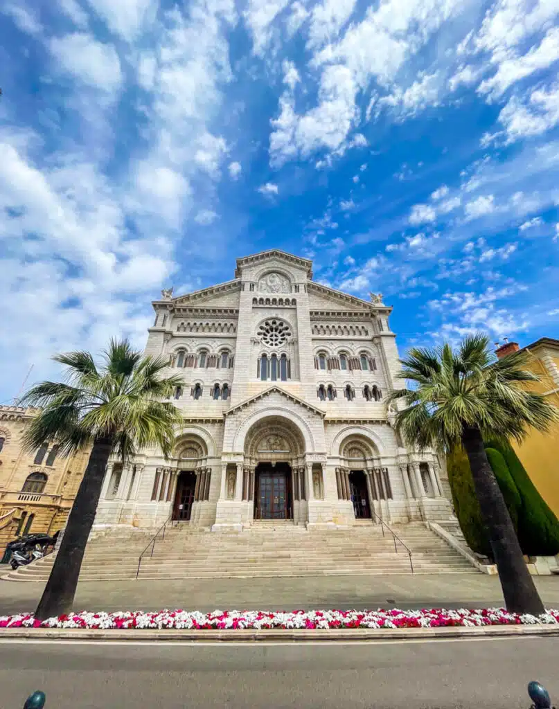 Front view of the white-stone Monaco Cathedral, framed by palm trees and colorful flowerbeds under a partly cloudy blue sky, captured during a day trip to Monaco from Nice.