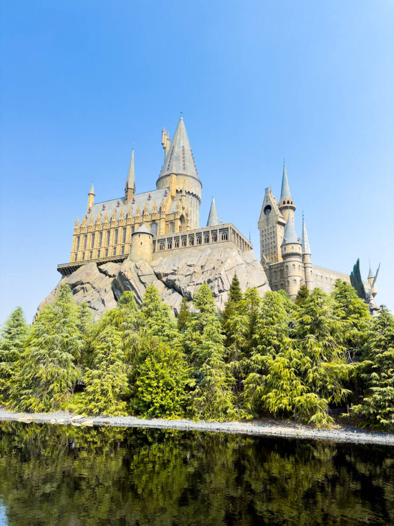 Majestic Hogwarts Castle attraction at Universal Studios Japan stands tall above a forested slope, reflected clearly in the water below under a bright blue sky.