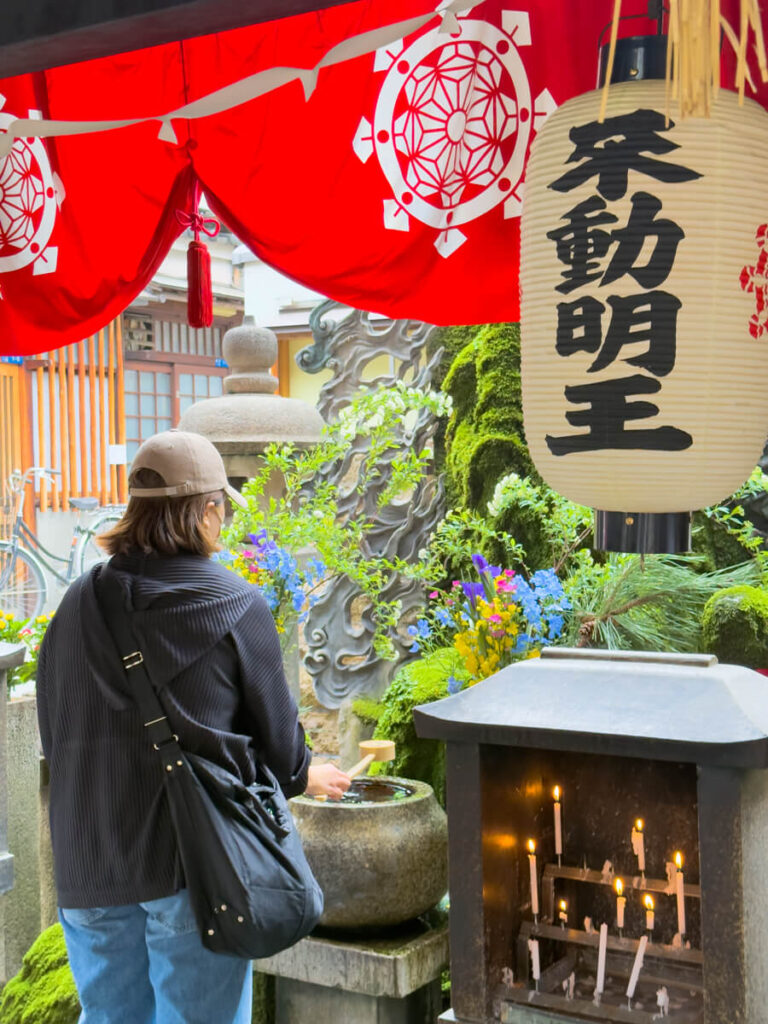 A woman in casual clothing stands at Hozen-ji shrine, using a ladle to scoop water from a stone basin surrounded by moss, flowers, and traditional lanterns with Japanese characters. A lit candle altar sits nearby under a vivid red curtain with white symbolic emblems.