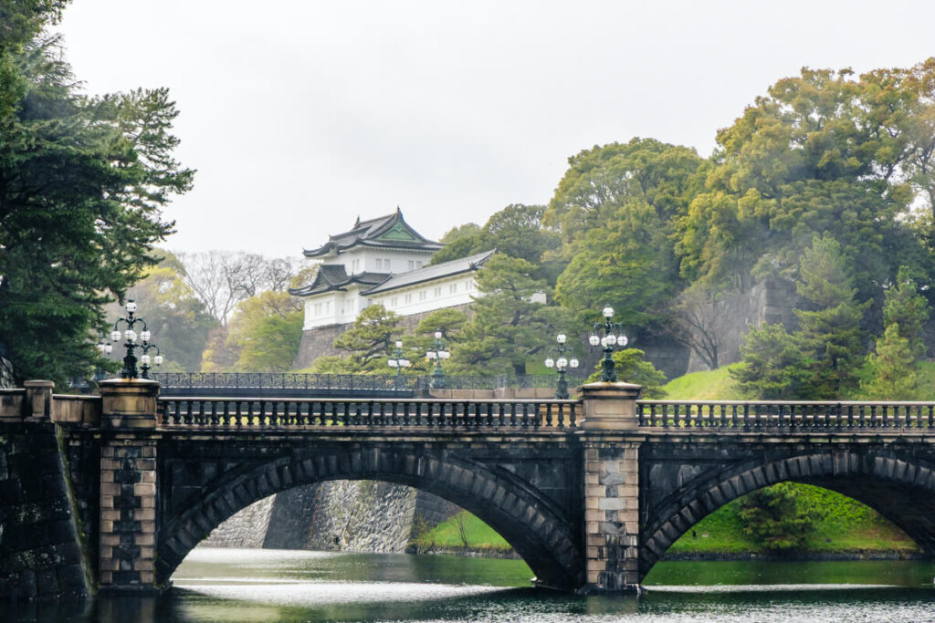 A misty view of Tokyo’s Imperial Palace with the iconic double-arched stone bridge, known as the Nijubashi Bridge, crossing the moat in the foreground. The white-walled palace building with a traditional green-tiled roof peeks through lush greenery, evoking a peaceful and regal atmosphere.