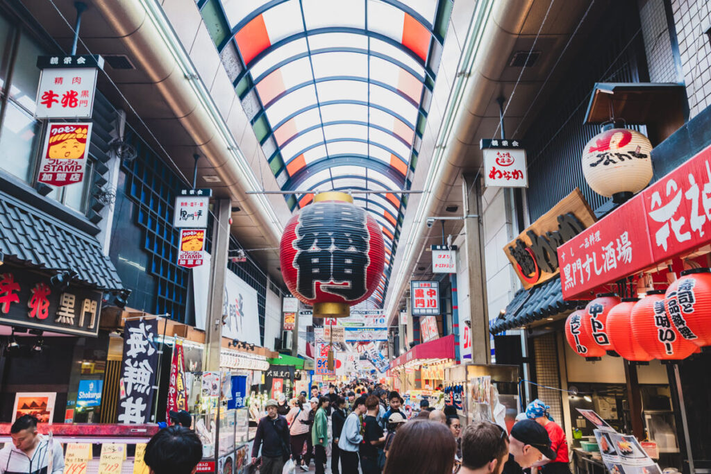 Crowded walkway inside Kuromon Ichiba Market in Osaka, with a large red paper lantern hanging from the ceiling and colorful vendor signs lining both sides. Shoppers explore food stalls under a curved glass roof with red, white, and green panels.