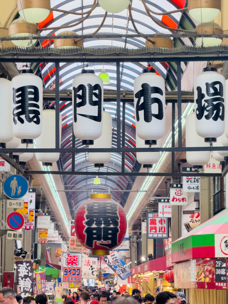 Large white and red lanterns with bold black kanji hang from the ceiling of the covered Kuromon Ichiba Market in Osaka, welcoming visitors to the bustling shopping arcade.