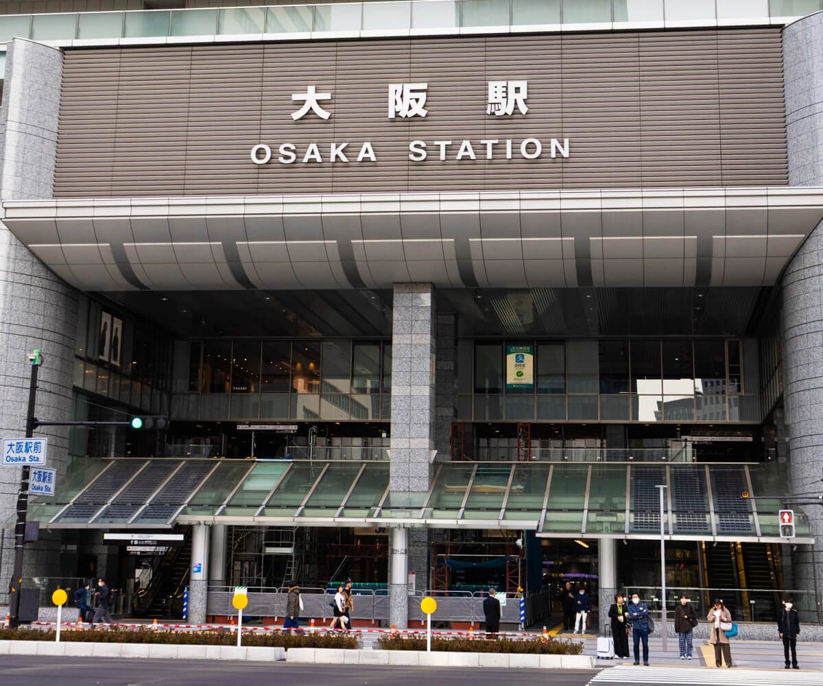 Front view of Osaka Station with its modern glass and stone exterior, large signage in Japanese and English, and people waiting at a crosswalk in the foreground. The entrance is flanked by escalators and a glass canopy, capturing the busy urban transit hub.