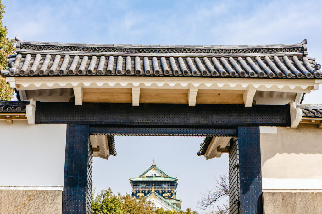 A distant view of Osaka Castle framed perfectly by a traditional Japanese gate in the foreground. The iconic green-roofed tower rises behind the black and white structure under a clear blue sky, showcasing classic Japanese architecture and historic significance.