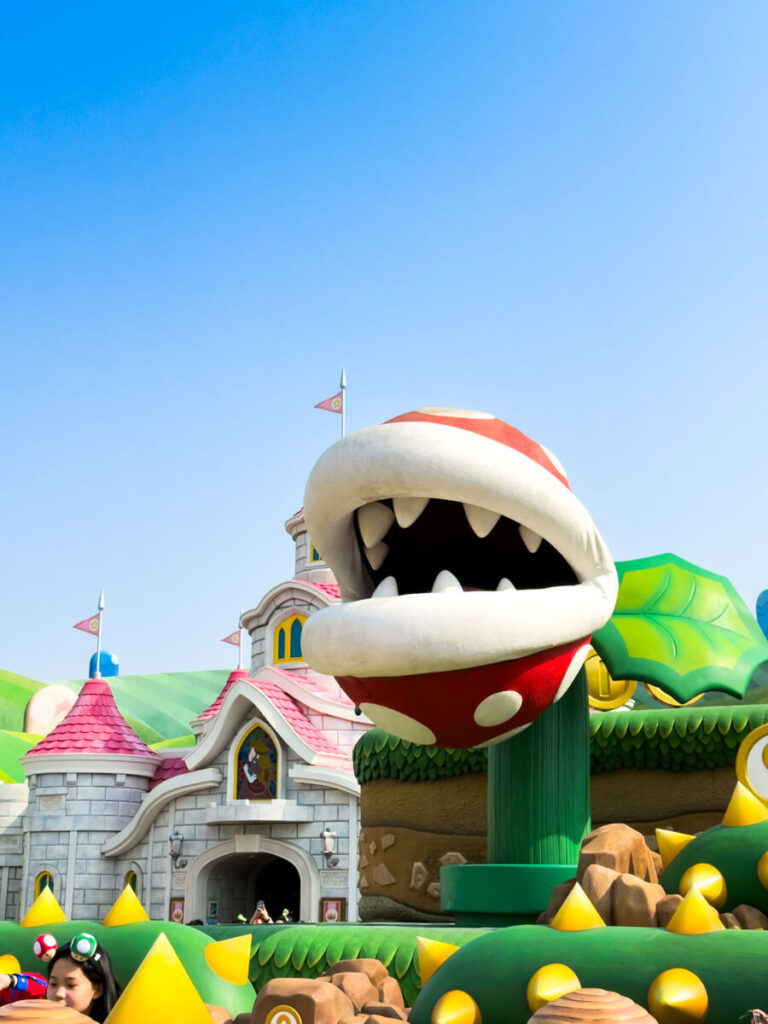 A large, animated Piranha Plant sculpture with sharp white teeth rises from a green pipe in Super Nintendo World, surrounded by vibrant themed landscaping. In the background, Princess Peach’s Castle features pink rooftops and playful flags under a bright blue sky.