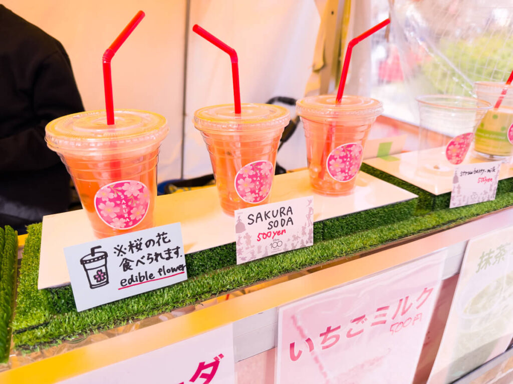 A row of plastic cups filled with pink Sakura soda sits on display at a festive Kyoto stall in Maruyama Park. Each drink includes edible cherry blossoms and is topped with a red straw, with handwritten signs in Japanese and English detailing the price and ingredients.