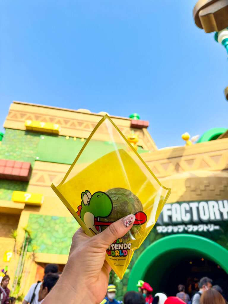 A hand holds up a green snack in Yoshi-themed packaging in front of Kinopio’s Café and the Toad Factory at Super Nintendo World.
