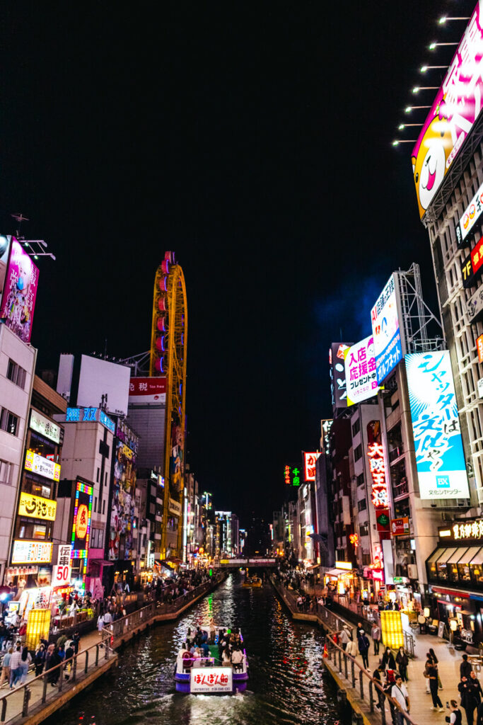 Night view of Dotonbori in Osaka with neon signs lighting up both sides of the canal and a sightseeing boat cruising down the water. The vibrant street scene includes the iconic yellow Ferris wheel on the Don Quijote building and crowds walking along the lively waterfront promenade.