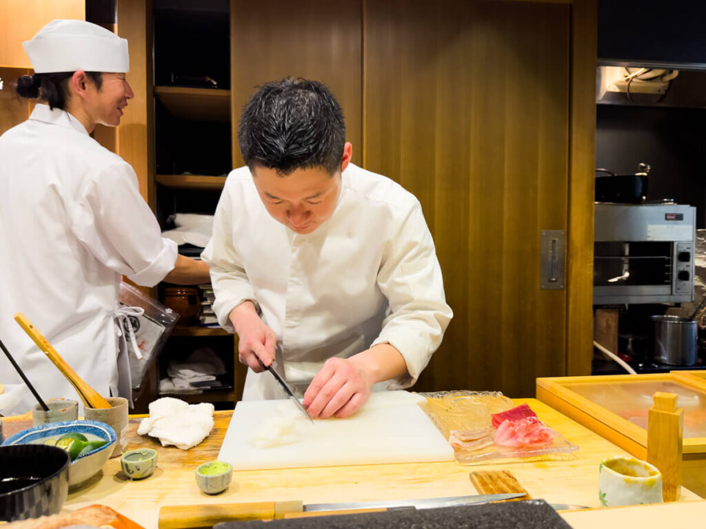 A chef carefully slices sashimi behind the counter during an omakase dinner in Osaka, Japan. The wooden counter is set with fresh fish, sauces, and small dishes, while another chef works in the background, adding to the intimate dining experience.
