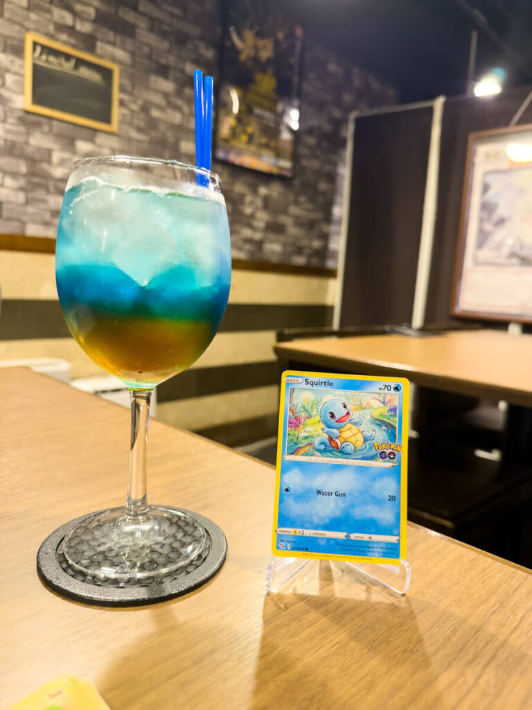 A layered blue and orange drink served in a wine glass next to a Squirtle Pokémon card on display at a themed café in Akihabara. The cozy interior and playful presentation reflect the area's love for anime and gaming.