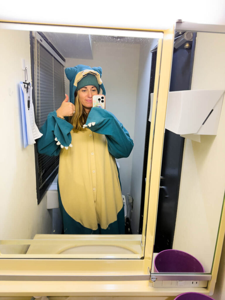 A woman poses in front of a mirror giving a thumbs up while wearing a cozy Snorlax onesie and beanie. She stands in a compact bathroom, capturing the playful and relaxed moment on her phone.