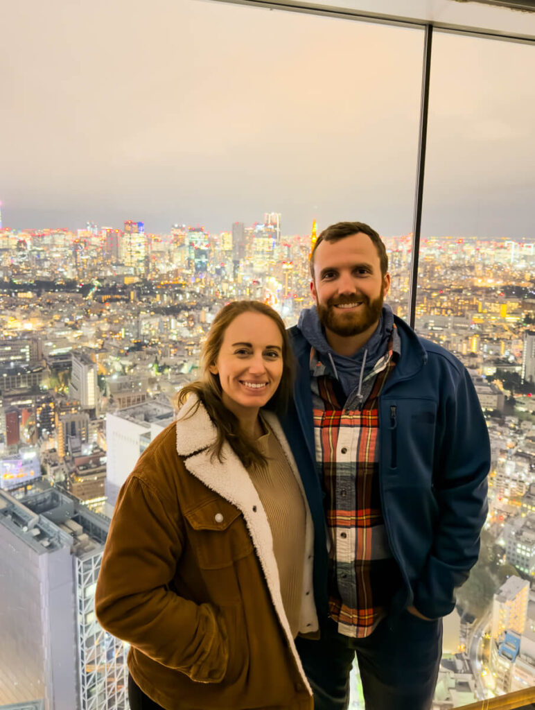 A couple poses in front of a glass window overlooking Tokyo’s dazzling city lights from Shibuya Sky observation deck. The sprawling metropolis sparkles beneath a dusky sky, capturing the vibrant energy of the city at night.