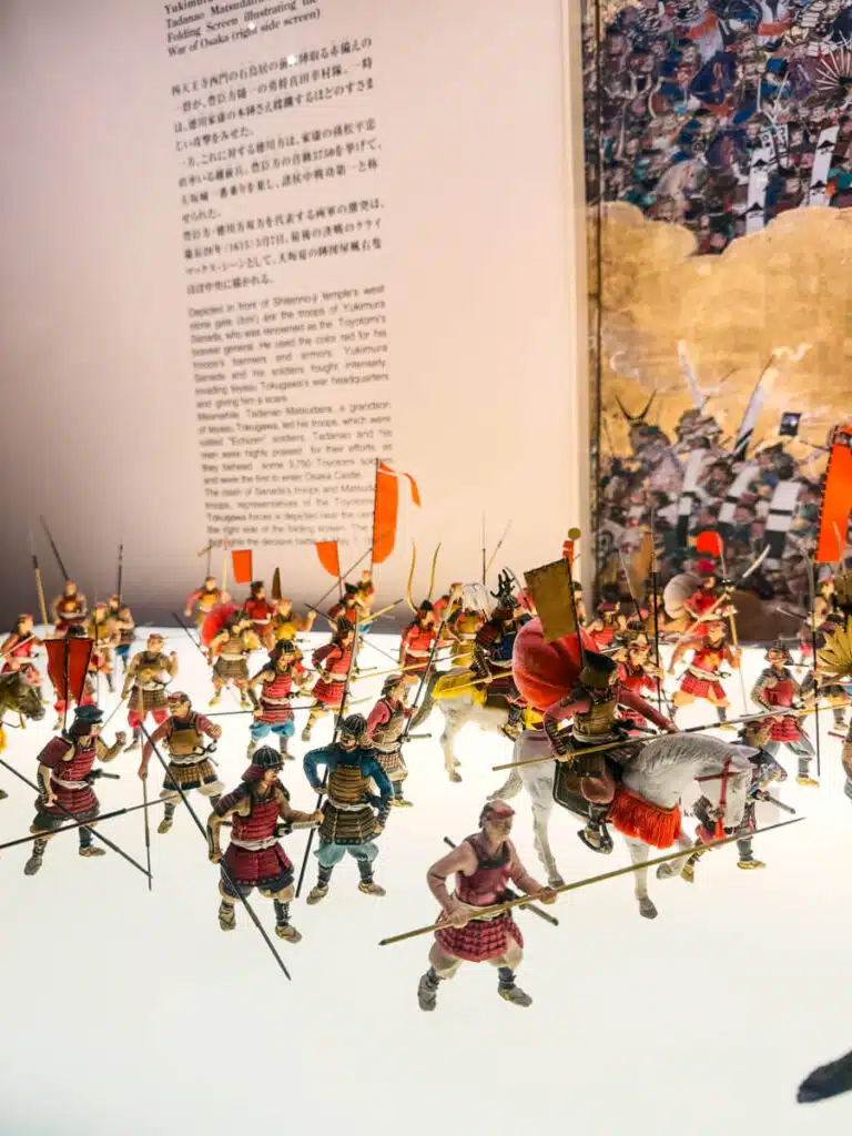 A detailed diorama of miniature samurai warriors in battle formation, part of an exhibit about the War of Osaka at the Osaka Castle Museum.