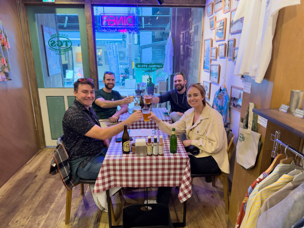 Four people sitting around a table with red-checkered cloth, toasting drinks and smiling inside Tagayasu General Store along Tenjinbashi-suji. The cozy diner-style setting features wood floors, framed photos on the wall, and a neon "Diner" sign glowing in the window.