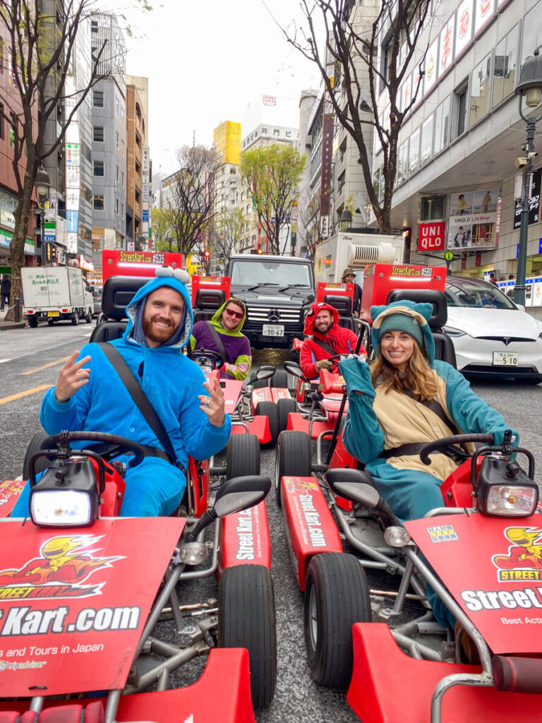 A group of friends dressed in colorful character costumes including Cookie Monster and Snorlax smile and pose in go-karts on a city street in Tokyo. The red “Street Kart” vehicles are lined up side-by-side, surrounded by tall buildings and bustling urban energy.