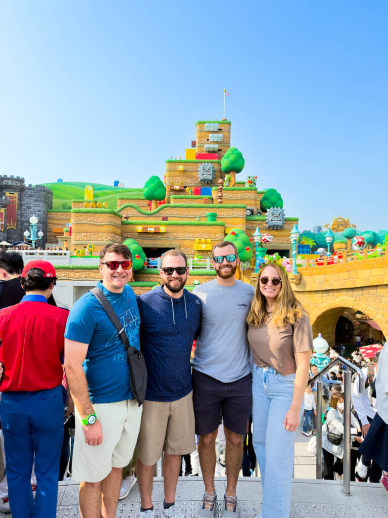 Four friends pose at the entrance of Super Nintendo World, with colorful blocks, green hills, and characters from the Mario universe in the background.
