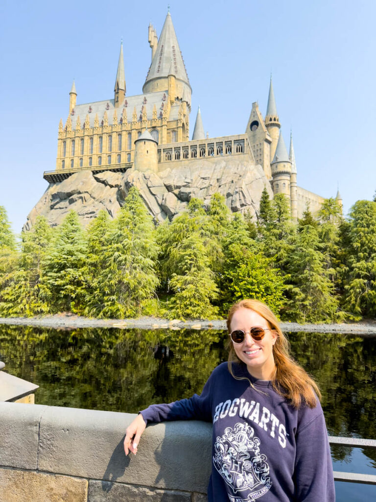 Woman wearing a Hogwarts sweatshirt smiles in front of the Hogwarts Castle replica at Universal Studios Japan, with the castle reflected in a calm, tree-lined pond behind her.