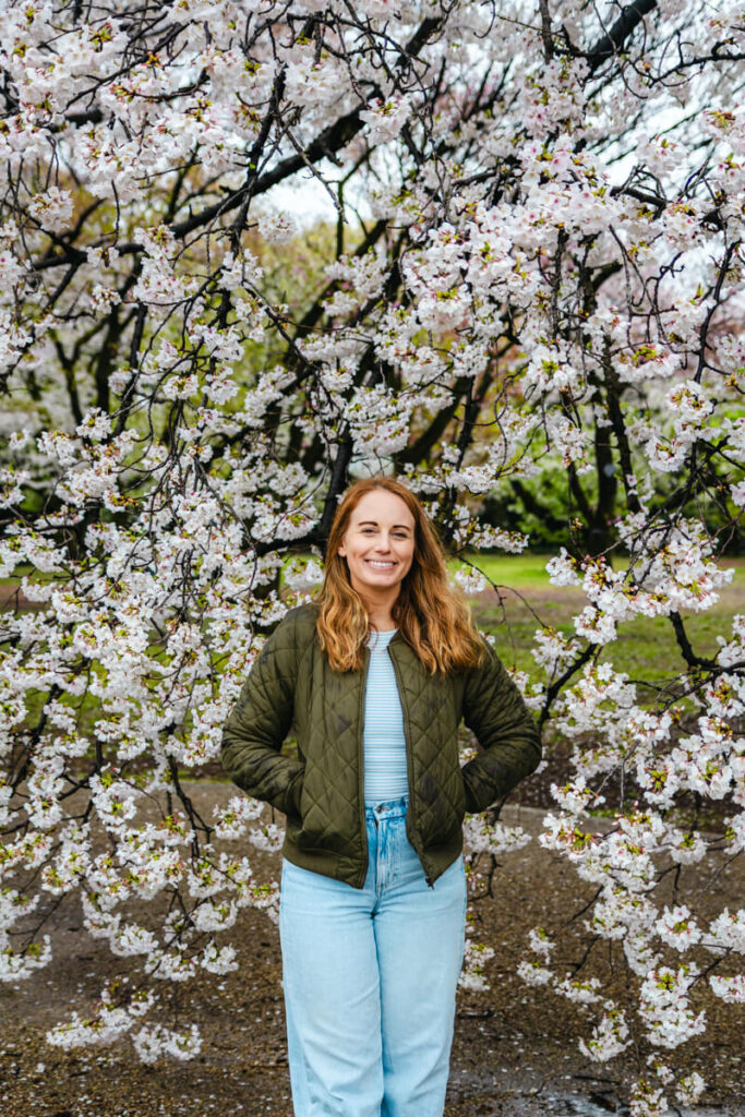 A woman in a green quilted jacket smiles in front of a tree full of white cherry blossoms in Shinjuku Gyoen National Garden. The delicate petals frame her as she stands with hands in her pockets on a damp path, capturing a peaceful spring moment.