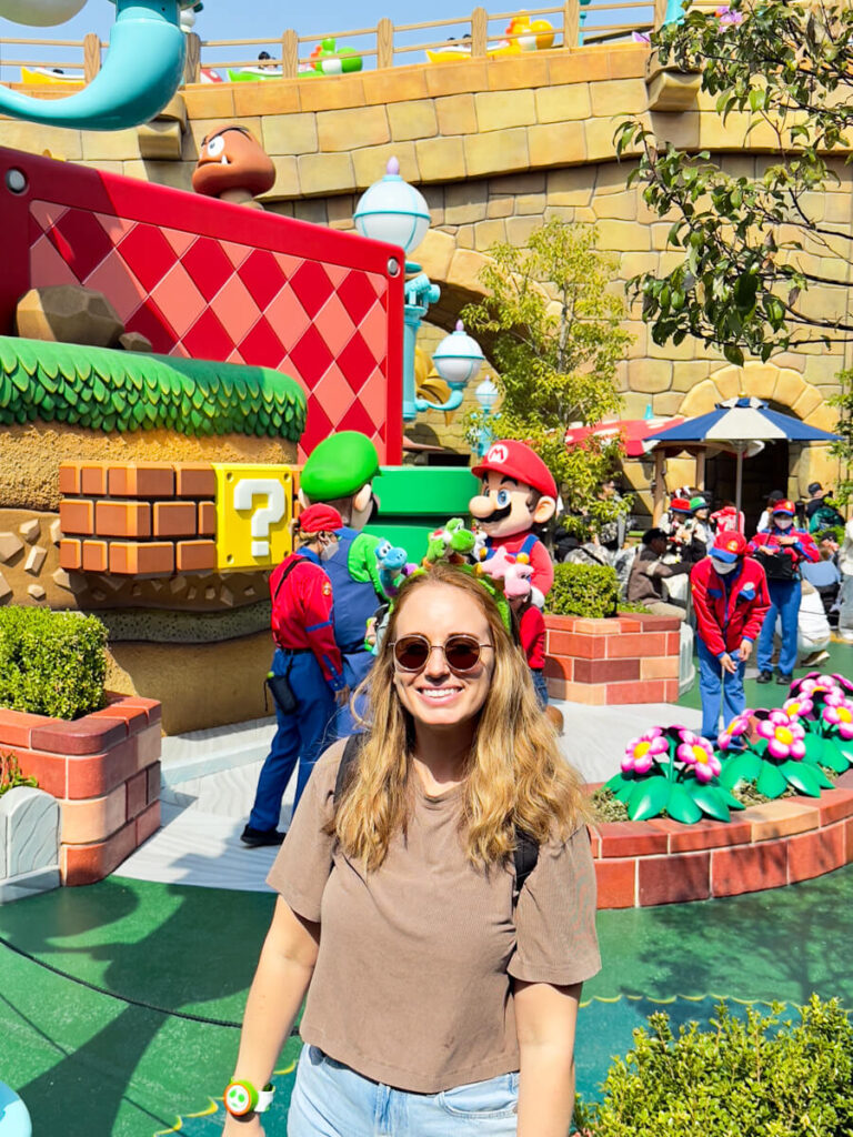 A woman stands smiling in front of life-size Mario and Luigi characters and vibrant themed scenery, including question blocks and colorful decor.
