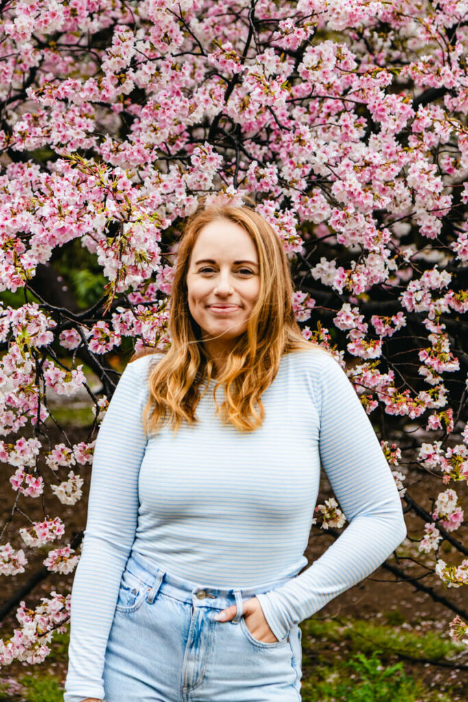 A woman in a light blue striped top smiles in front of a blooming cherry blossom tree in Shinjuku Gyoen National Garden. The branches are densely covered in soft pink sakura flowers, creating a vibrant spring backdrop.