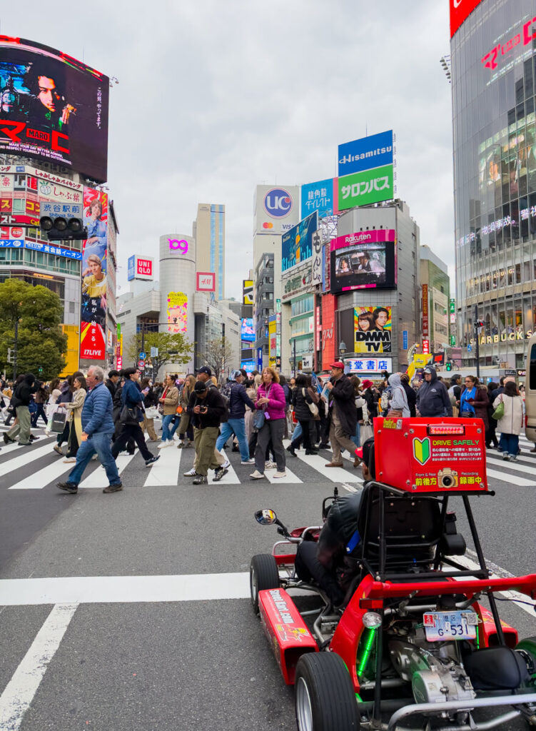 A red go-kart driver waits at the iconic Shibuya Crossing in Tokyo as a wave of pedestrians cross the street. Towering buildings and massive digital billboards fill the background in one of the world’s busiest intersections.