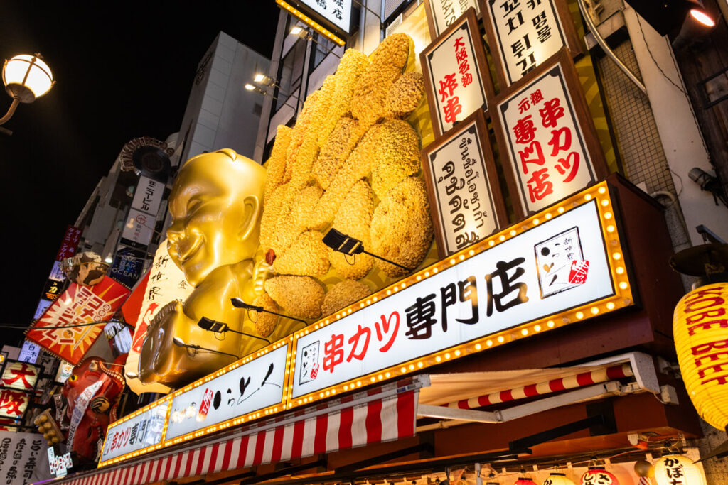 A glowing kushikatsu restaurant in Osaka's Shinsekai district features a large golden statue of Billiken and oversized fried food models lit up under neon signs.