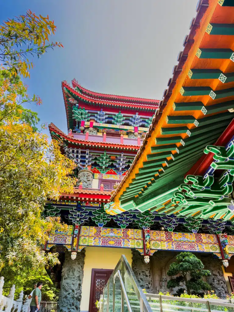 Upward view of Po Lin Monastery’s colorful pagoda and rooflines, highlighting the detailed ornamentation and vivid paintwork. Trees and greenery frame the edge of the temple, adding to its serene setting.