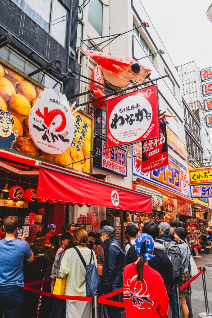 A crowd lines up outside a popular takoyaki shop in Osaka featuring bright red signage, cartoon-style branding, and a large octopus sculpture above the storefront.