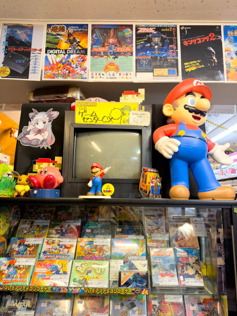 A colorful retro game shop display in Akihabara, Tokyo, featuring a vintage CRT TV, large Super Mario statue, and shelves full of classic Famicom game cartridges and box art. Posters and anime stickers decorate the wall above, capturing the nostalgic charm of Japanese gaming culture.