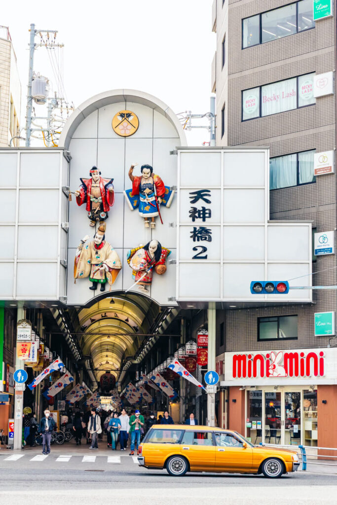 Entrance to Tenjinbashi-suji shopping street featuring large, colorful kabuki-style figures mounted above the entryway between two buildings. Below, people walk through the bustling arcade while a retro yellow station wagon passes by on the street.