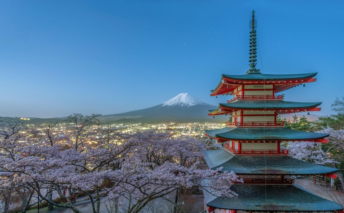 A breathtaking view of Chureito Pagoda framed by cherry blossoms in full bloom, with the iconic snow-capped Mount Fuji in the background. The city lights of Fujiyoshida shimmer below under a twilight sky, capturing a serene and iconic scene of springtime in Japan.