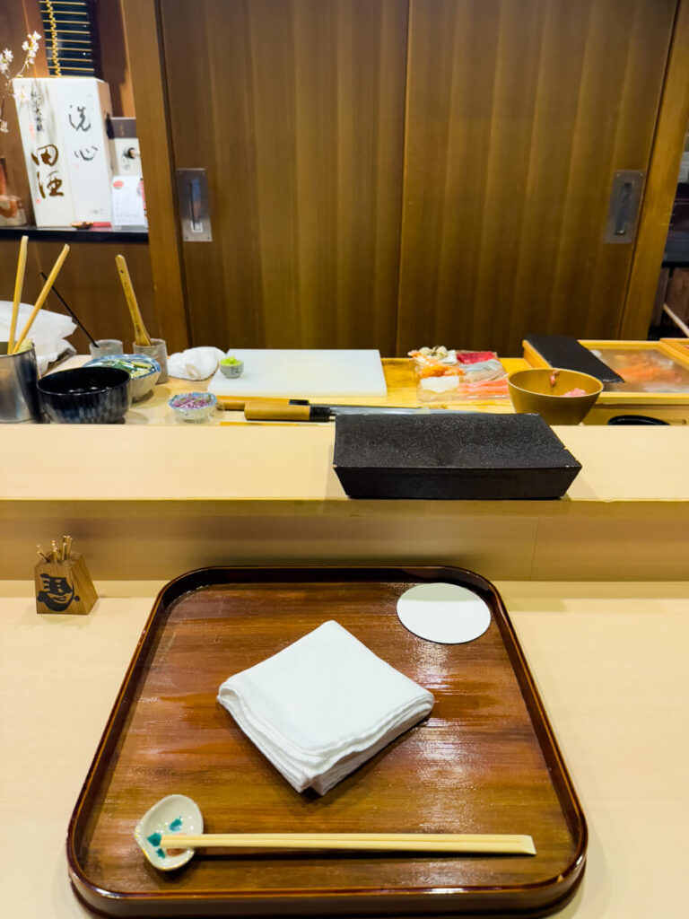 Minimalist sushi counter setting in Japan with a wooden tray holding chopsticks, a folded white napkin, and a ceramic chopstick rest. Behind the counter are sushi ingredients, knives, and preparation tools arranged neatly on a wooden board, ready for omakase service.