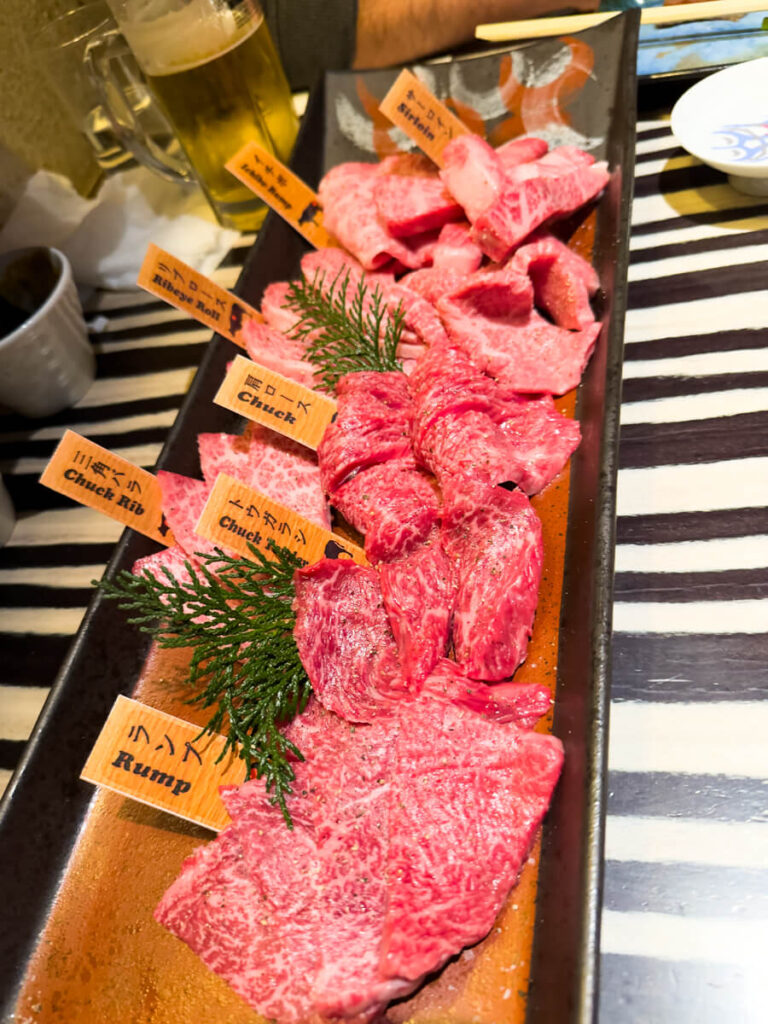 A beautifully arranged platter of raw Wagyu beef slices labeled with cut names such as "Chuck Rib," "Ribeye Roll," and "Rump," ready for a Japanese BBQ (Yakiniku) meal. A cold glass of beer sits in the background on a striped tabletop.