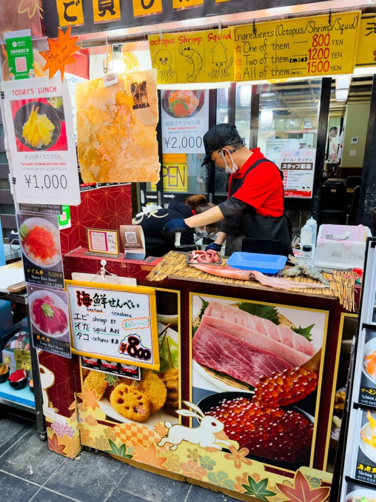 A vendor prepares fresh seafood crackers made with shrimp, squid, and octopus at a colorful food stall adorned with photos, drawings, and lunch set menus.