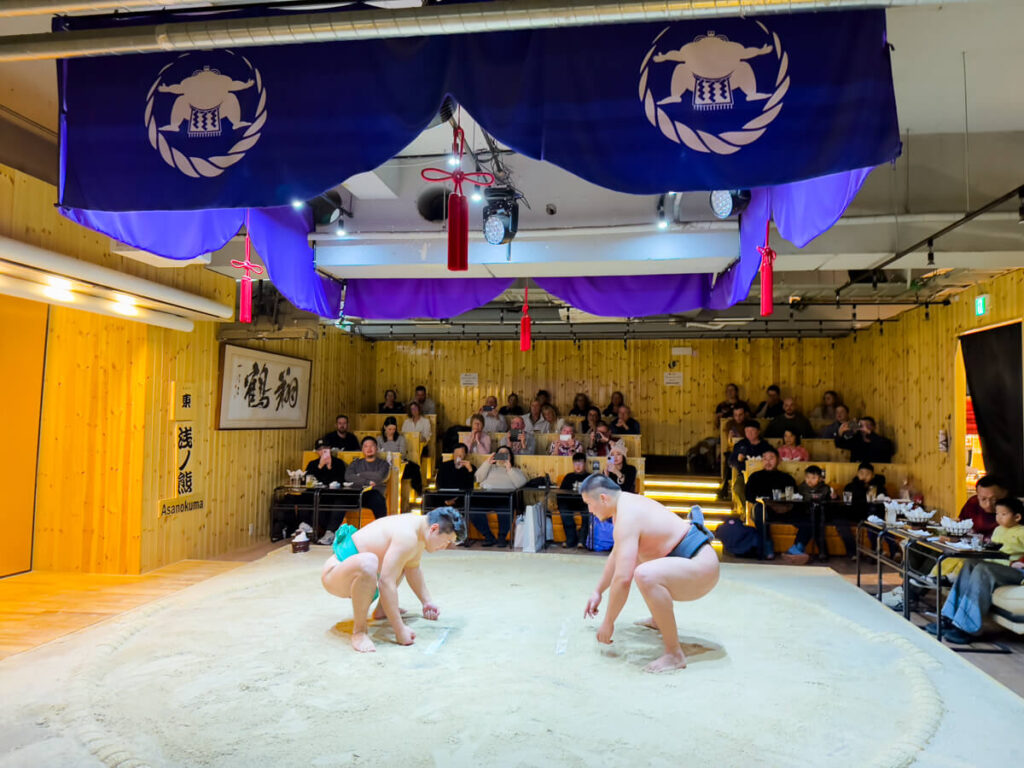 Two sumo wrestlers crouch in a ready stance inside a circular dohyo ring during a live match at a sumo-themed restaurant in Tokyo. The wooden interior, seated diners, and purple banners overhead create an intimate yet theatrical atmosphere where guests enjoy food while watching traditional Japanese wrestling.