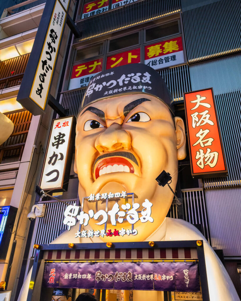 Eye-catching storefront on Dotonbori Street in Osaka featuring a giant, scowling chef’s face as the facade of the famous Kushikatsu Daruma restaurant. The bold signage and illuminated lights draw attention to this well-known spot for deep-fried skewers.