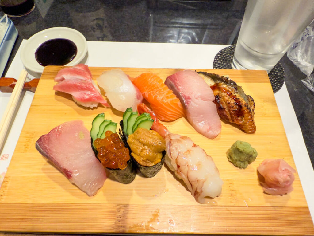 A variety of fresh sushi, including tuna, salmon, eel, uni, and ikura, is served on a wooden tray with wasabi, pickled ginger, and soy sauce on the side.