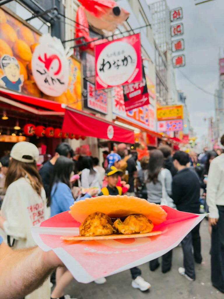 A close-up of two takoyaki balls served in a crispy shrimp cracker is held up in front of a busy street food stall in Osaka with red signage and a crab sculpture above.