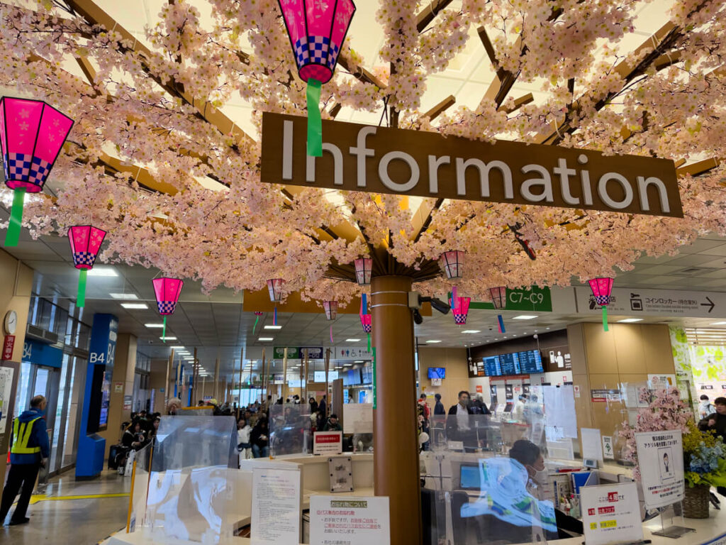 Airport information desk beneath artificial cherry blossom branches decorated with pink lanterns and a large hanging Information sign. Travelers gather around the counter while staff assist visitors in a bright terminal decorated for spring.