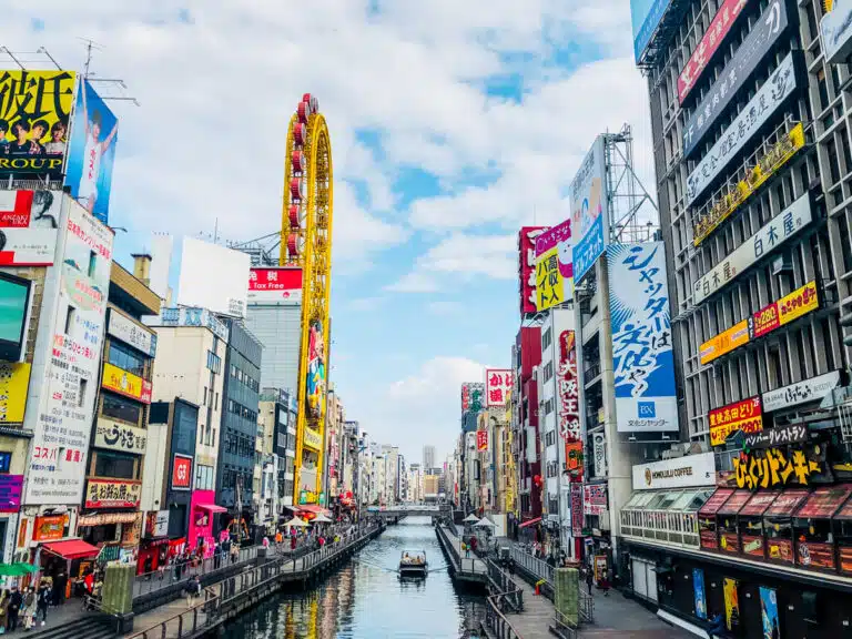 A daytime view of Dotonbori in Osaka, Japan, featuring the lively canal lined with colorful billboards, busy shops, and the iconic yellow Ferris wheel attached to the Don Quijote building. The image captures the energetic atmosphere perfect for any Osaka itinerary.
