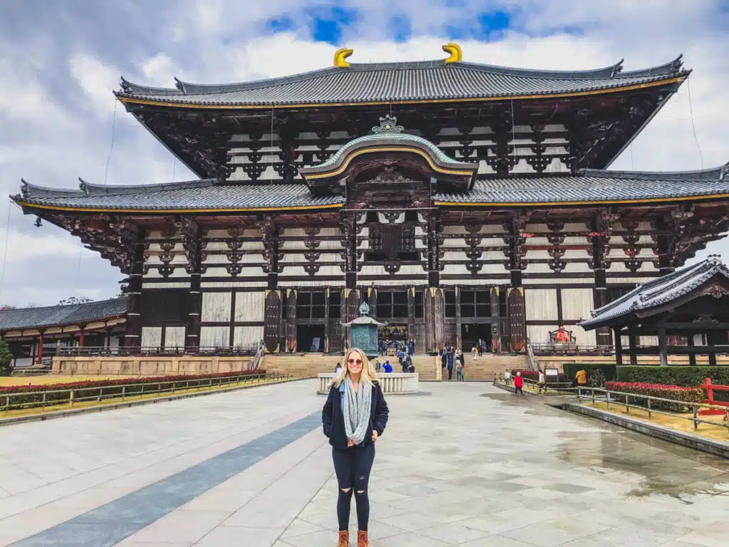 A woman stands smiling in front of the massive Great Buddha Hall of Todai-ji Temple in Nara, showcasing the grandeur of the wooden architecture.