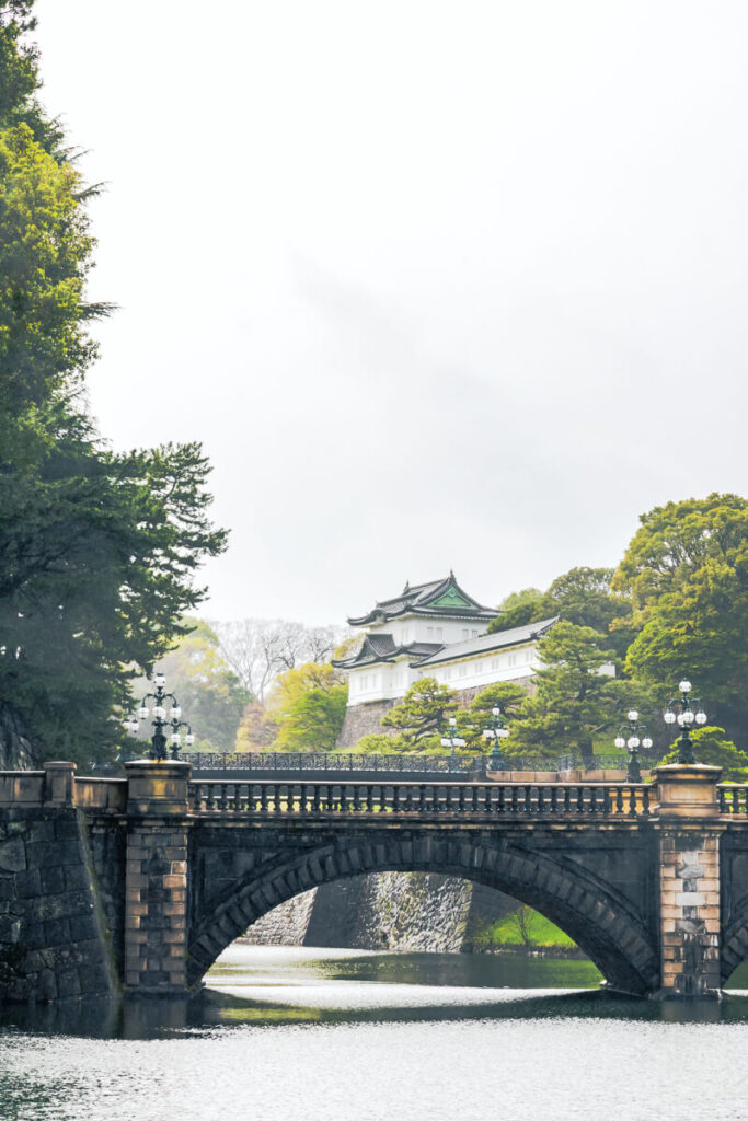 Stone bridge arching over a calm moat with the Imperial castle and green tiled roof visible beyond the trees. The historic structure sits behind fortified stone walls surrounded by lush greenery in central Tokyo.