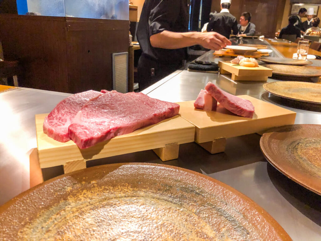 Raw Kobe beef steaks with heavy marbling displayed on wooden blocks at a teppanyaki counter in Kobe Japan while a chef prepares the grill in the background.