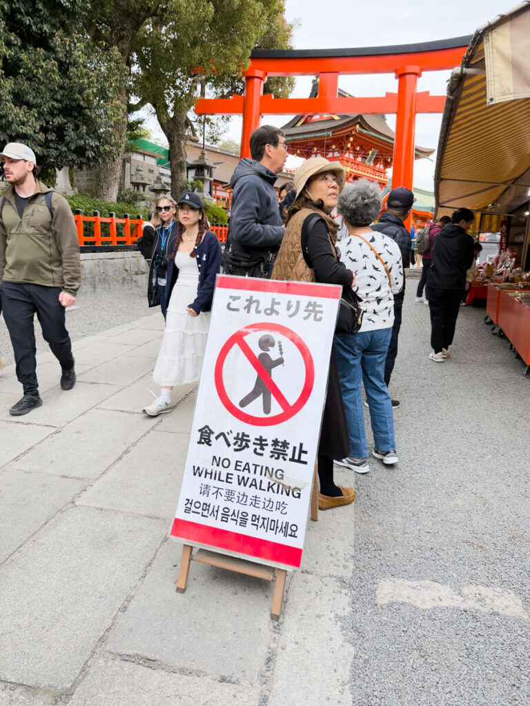 Outdoor walkway near a shrine with a bright red torii gate in the background and market stalls on the right. A standing sign in the foreground reads “No Eating While Walking” in multiple languages. Several tourists are gathered around the entrance area.