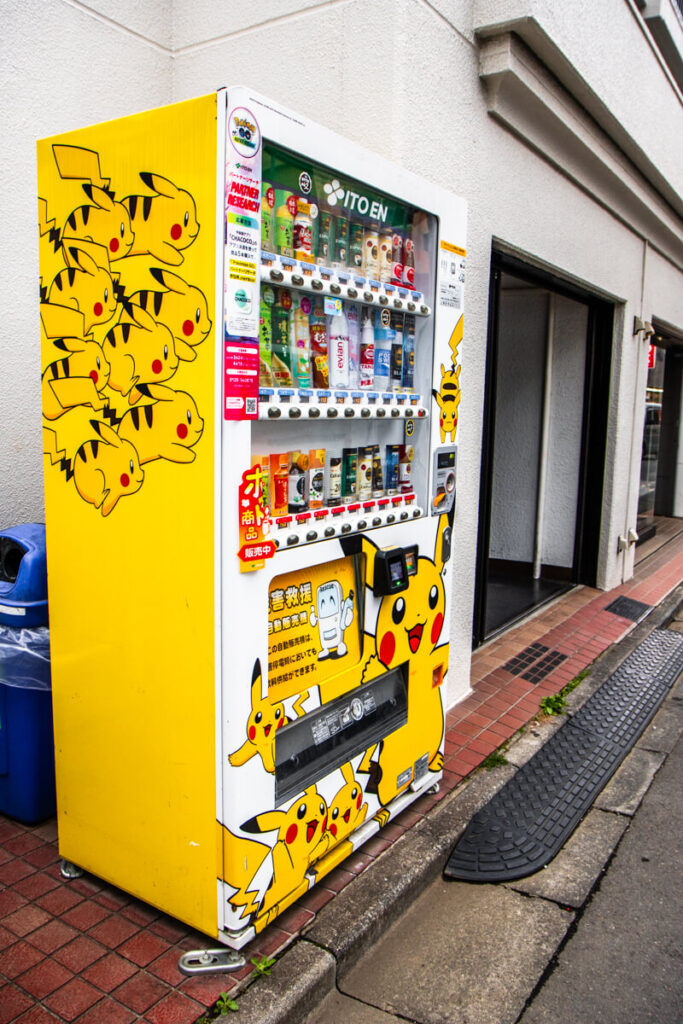 Bright yellow vending machine decorated with multiple Pikachu characters selling bottled drinks on a sidewalk in Japan. The colorful Pokemon themed machine stands against a white building wall attracting attention from passersby.