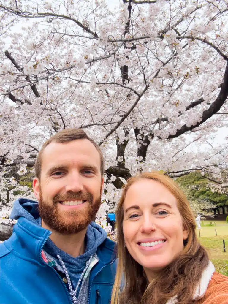 A smiling couple takes a selfie under full-bloom cherry trees in Yoyogi Park, capturing a joyful moment during sakura season in Japan.