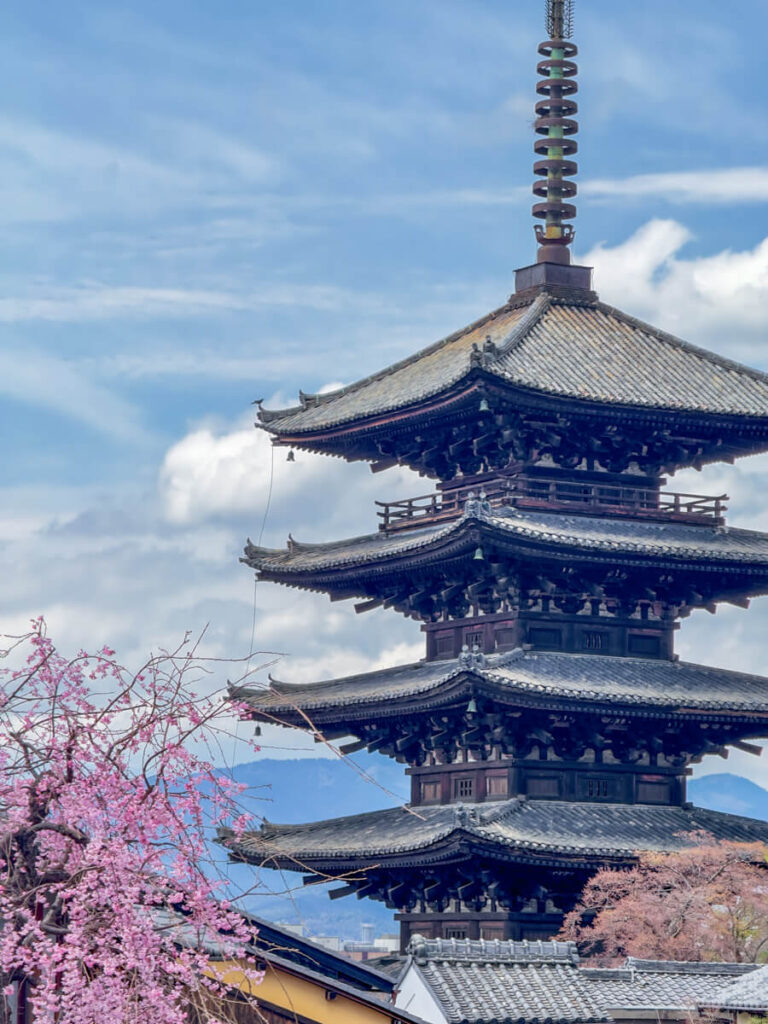 Yasaka Pagoda looming in front of a cloudy blue sky with a cherry blossom tree blooming in the left corner